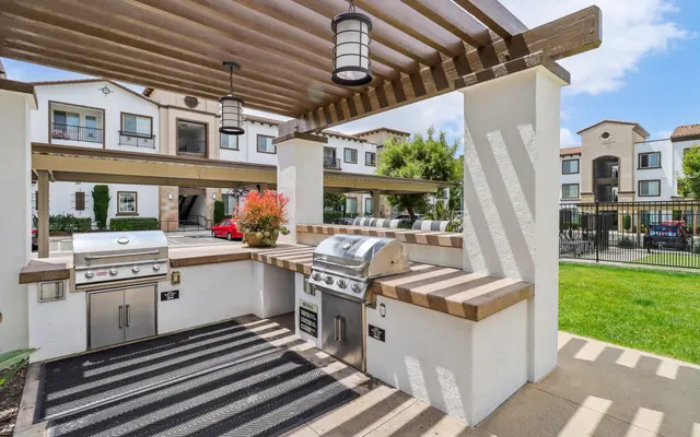 A well-designed outdoor grilling area featuring a barbecue grill and food prep space under a pergola, surrounded by greenery and residential buildings in the background.