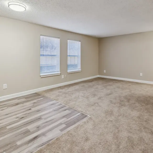 An empty living room with two windows, beige walls, and a mix of carpet and laminate flooring.