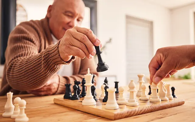 Strategic Chess Game Two people playing chess at a wooden table, with one person moving a black piece and the other adjusting a white piece.