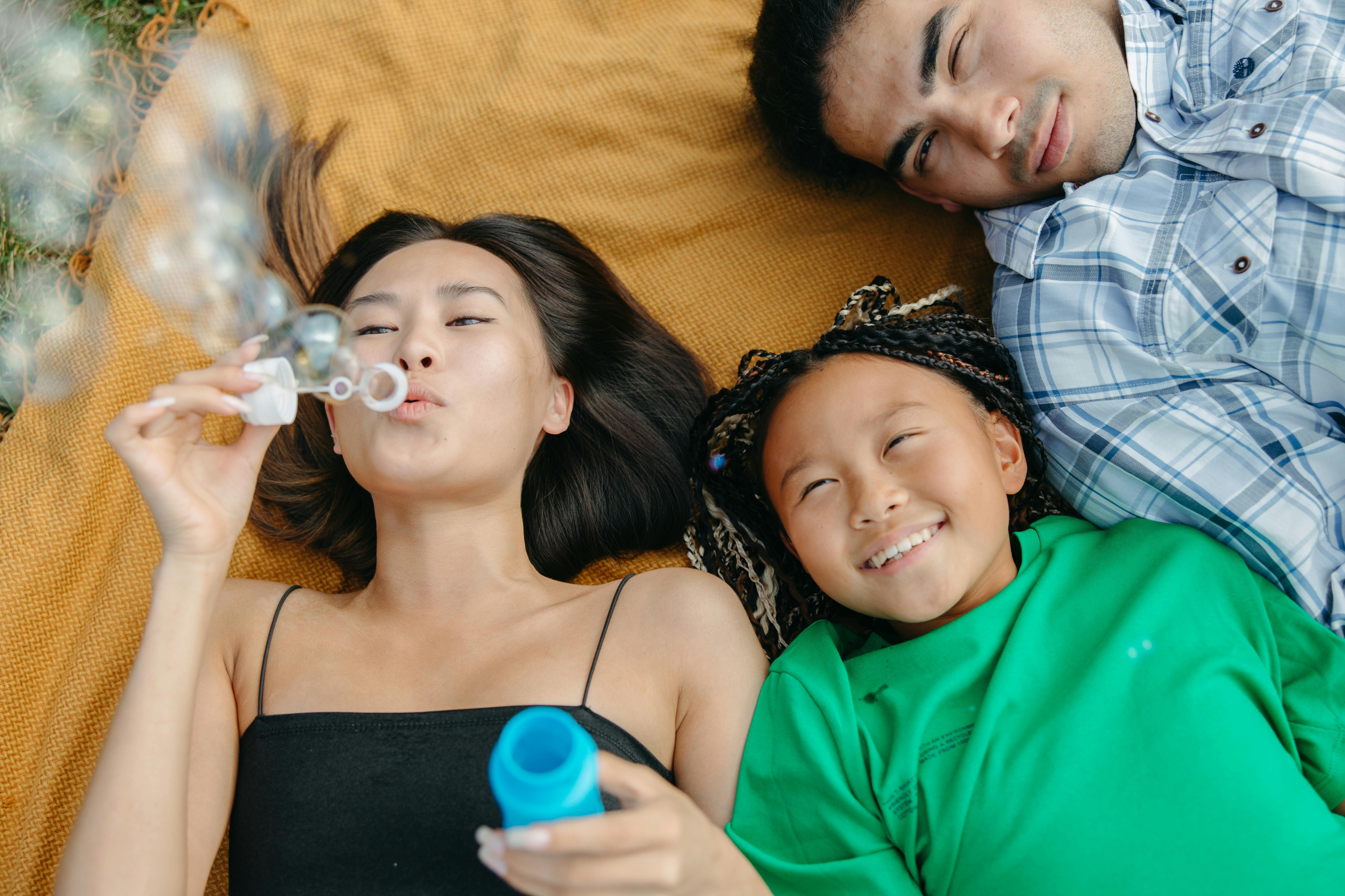 A woman blowing bubbles while a girl smiles beside her, and a man rests nearby, all lying on a blanket outdoors.