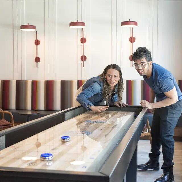 Two people playing a shuffleboard game in a modern lounge area with wooden decor and colorful seating.