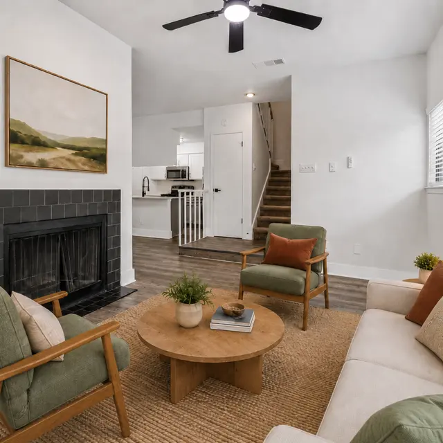 A modern living room featuring a cozy seating area with a white sofa, two green armchairs, and a round wooden coffee table. A black fireplace is beneath a landscape painting on a white wall, with a view of a kitchen and stairs in the background.