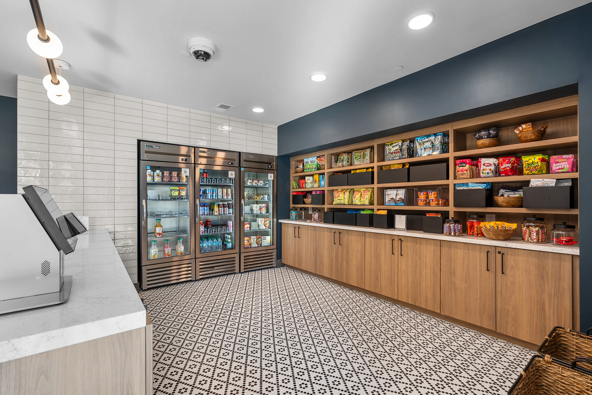 A modern convenience store interior featuring a refrigerated display case, wooden shelves stocked with various snacks, and a stylish patterned floor.