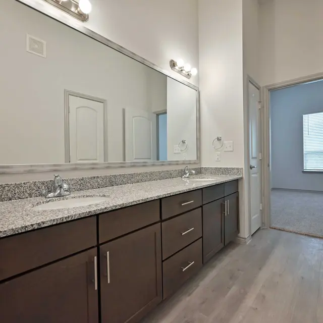 A modern bathroom featuring a double vanity with a long countertop and a large mirror. The cabinetry is dark wood, and the countertop is speckled grey. To the right, there is a doorway leading to another room with a window.
