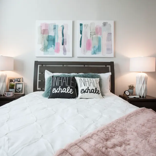 A modern bedroom featuring a large bed with white comforter and decorative pillows that read 'INHALE' and 'exhale'. The room is softly lit with two bedside lamps on dark wooden nightstands, and abstract art pieces are hung above the bed. A cozy pink throw blanket is draped over the corner of the bed.