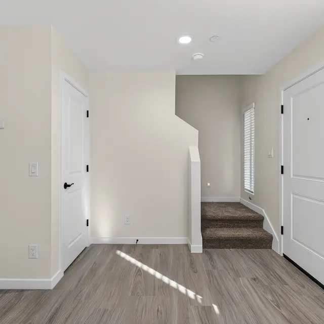 A spacious entryway with light-colored walls, two closed doors on the left and a staircase leading up on the right. Large window allows natural light. Wooden floor.