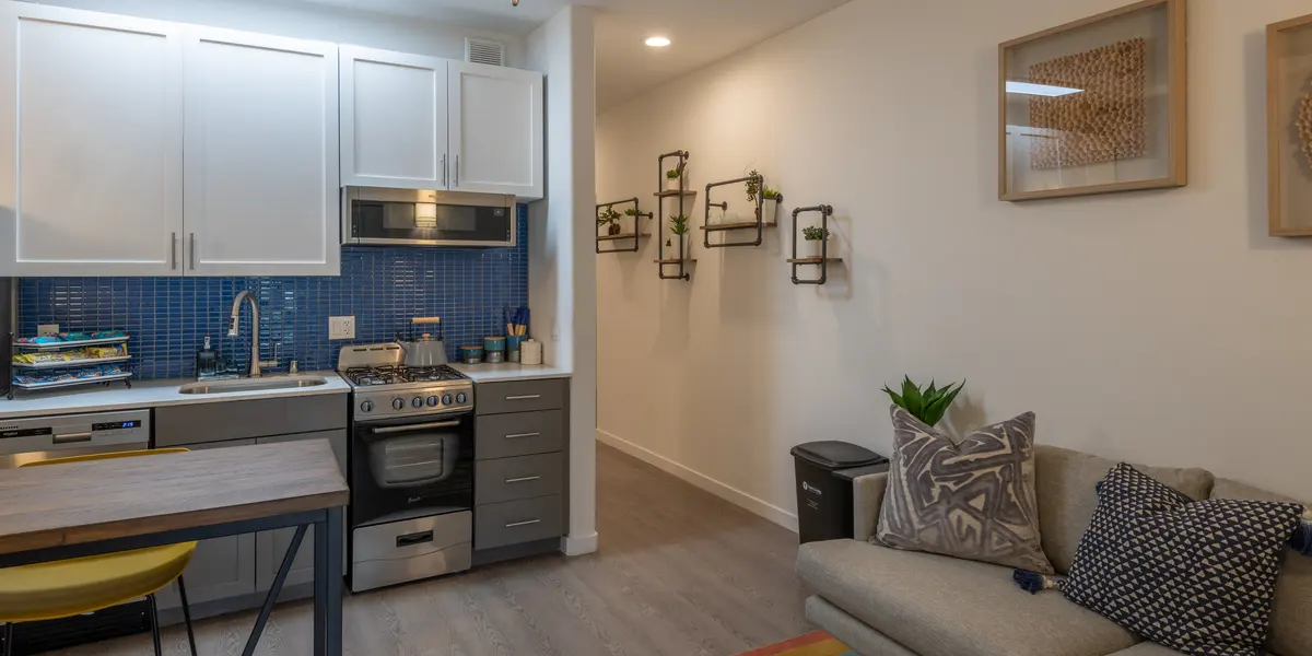 A modern kitchen and living area featuring a stove, a small dining table, and a couch with decorative pillows. There are blue tiles on the backsplash and some decorative wall shelves. A colorful rug is visible on the floor.