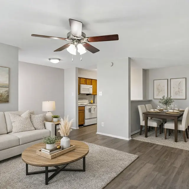 Cozy Modern Living Space A spacious living area featuring a light gray couch, a round wooden coffee table, and a ceiling fan. Adjacent is a dining area with a table and chairs, and a view of a kitchen in the background. The walls are painted in soft colors, and there are decorative elements like artwork and plants.
