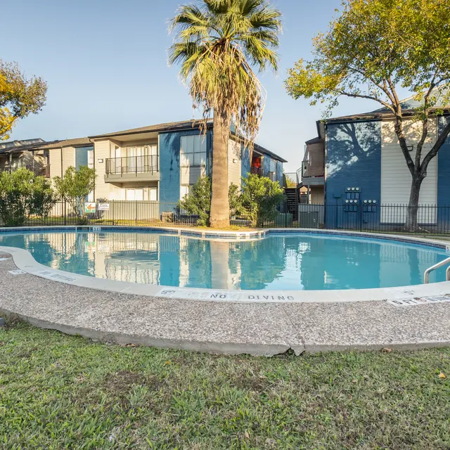 View of an outdoor swimming pool surrounded by grass and trees, with apartment buildings in the background.