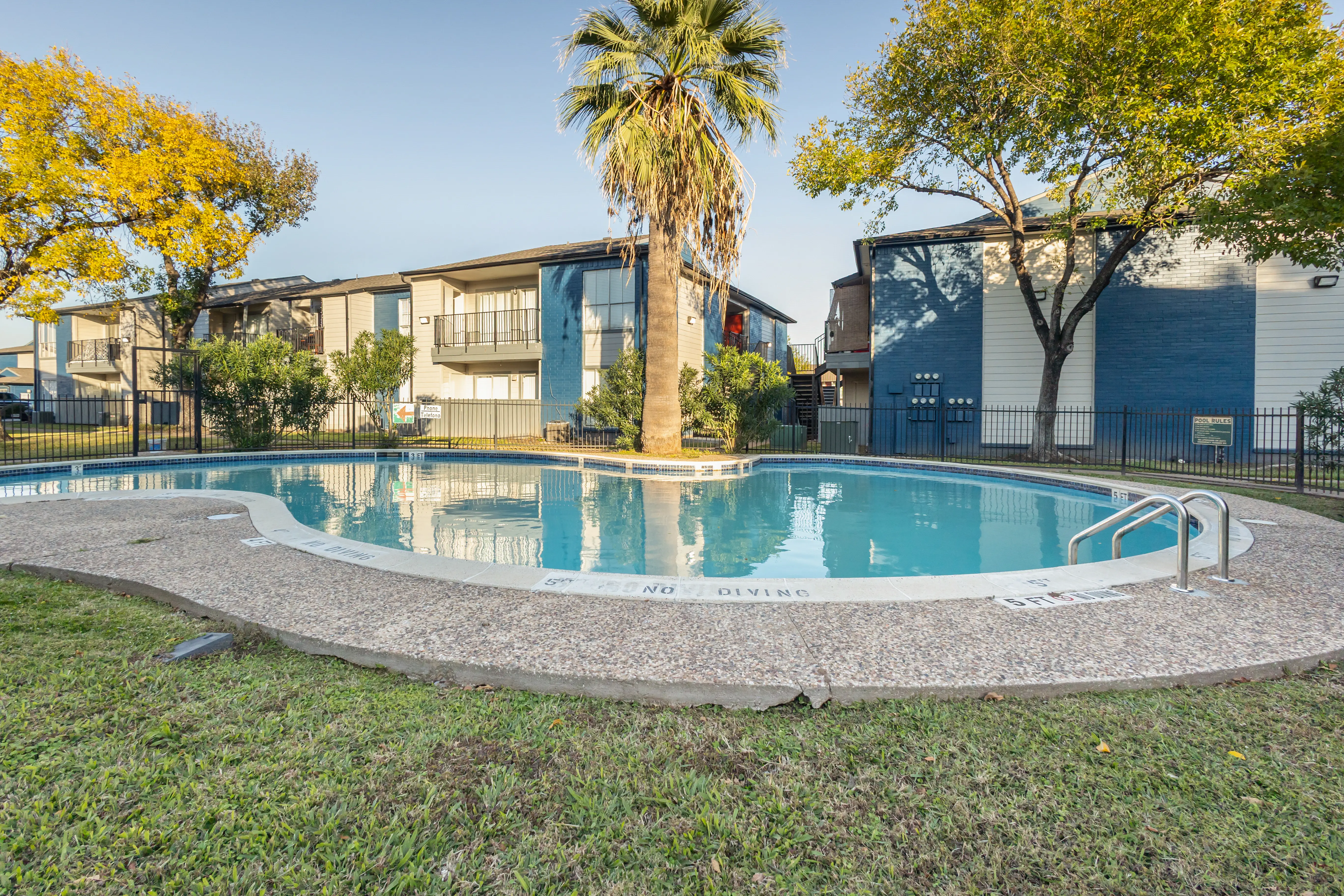 View of an outdoor swimming pool surrounded by grass and trees, with apartment buildings in the background.