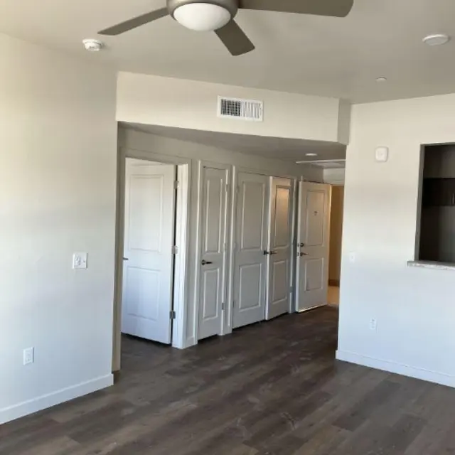 A view of a modern apartment interior featuring a ceiling fan, light-colored walls, and wooden floors. In the background, several closed doors are visible. The overall design is clean and minimalistic.