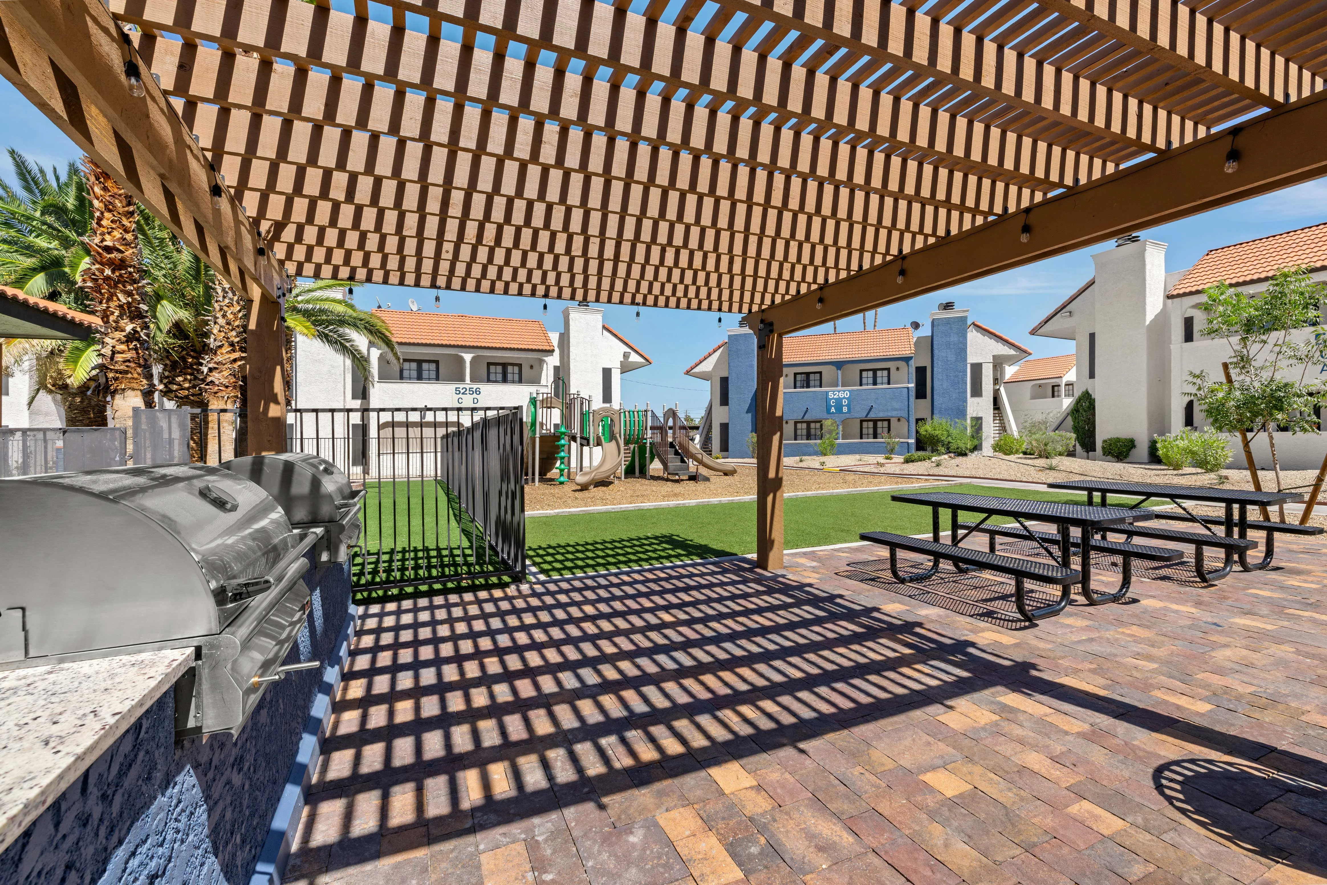 An outdoor barbecue area featuring grills, picnic tables, and a playground visible in the background, surrounded by residential buildings and palm trees.