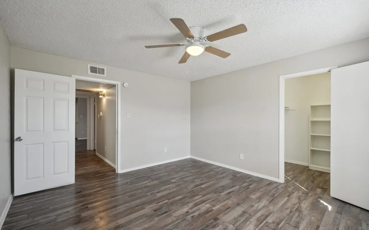 Empty bedroom with a ceiling fan and wooden flooring, featuring two open doors leading to adjacent rooms.