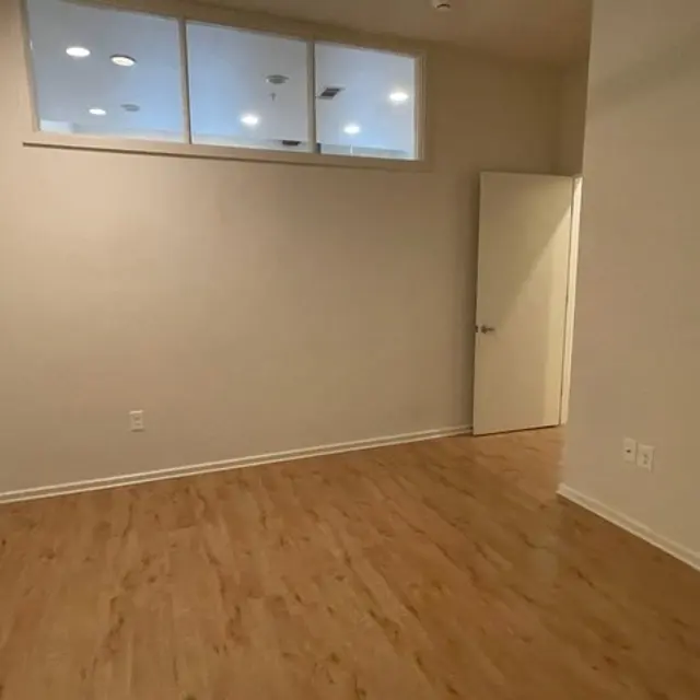 A vacant room with light wooden flooring and beige walls. The room features a door on the right and a small window near the ceiling that allows light in.