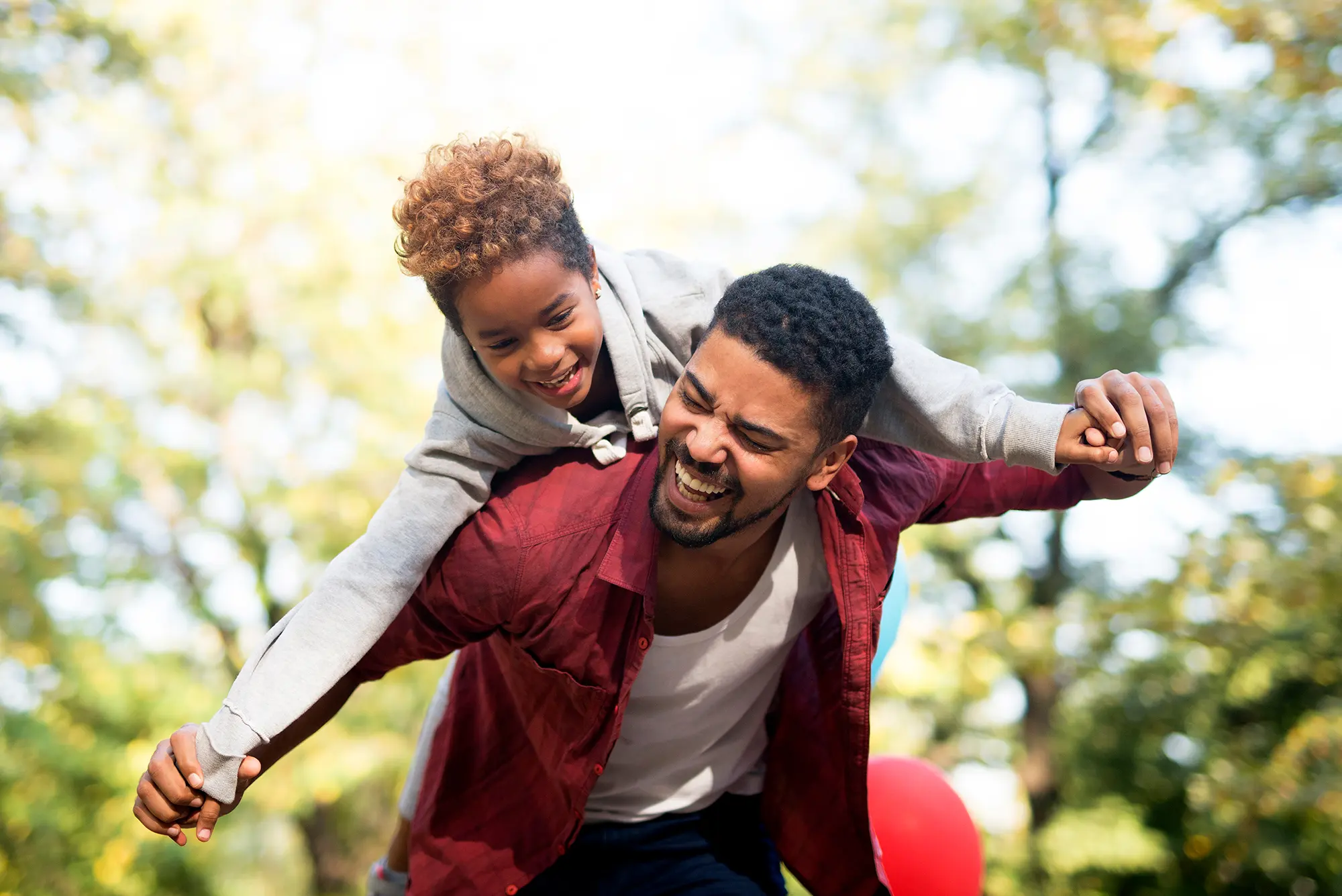 A father carries his son on his back, both smiling and enjoying a sunny day outdoors surrounded by greenery.