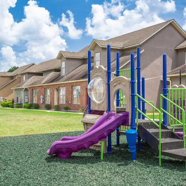 A vibrant playground with a purple slide and green climbing structure surrounded by residential buildings and lush grass under a bright blue sky with fluffy clouds.