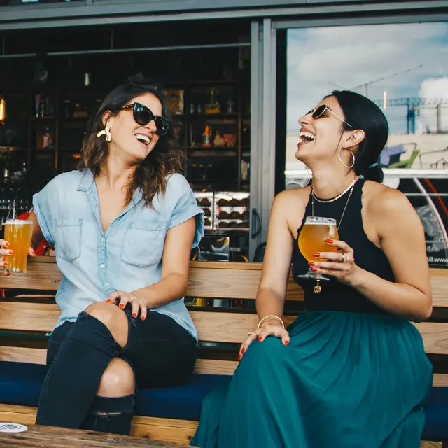 Laughing Friends at the Bar Two women sitting on a bench laughing and enjoying drinks at a bar. One woman is wearing a light blue shirt with sunglasses, and the other is dressed in a black top and green skirt.