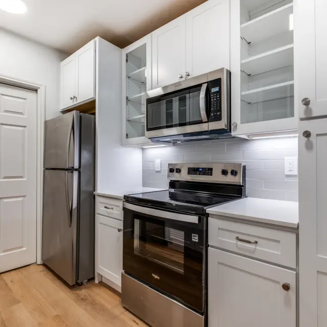 A modern kitchen featuring white cabinets, a stainless steel refrigerator, a microwave, and an oven. The countertops are light-colored and the floor is wooden.