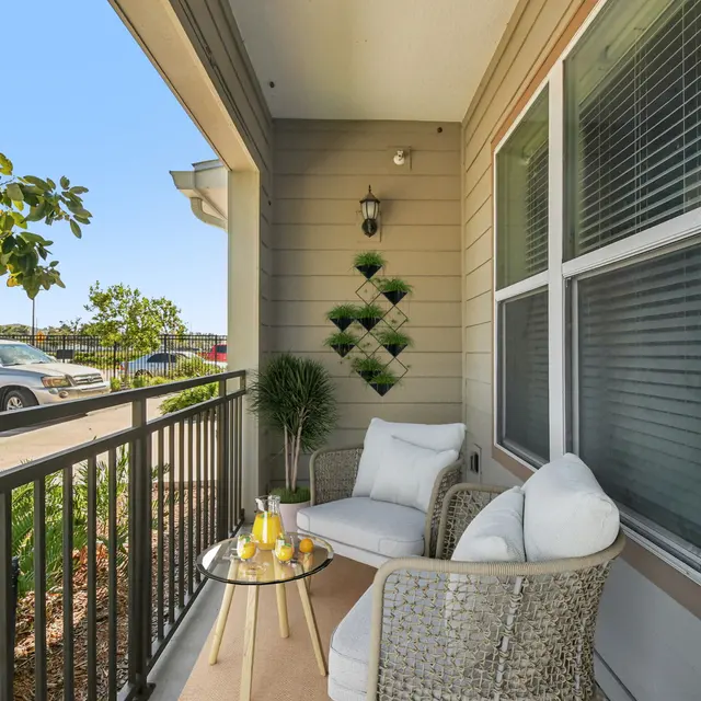 A cozy balcony featuring two white chairs with a small glass table. A decorative plant is on the table, and a wall-mounted plant display is in the background. Facing greenery and parked cars are visible in the background.