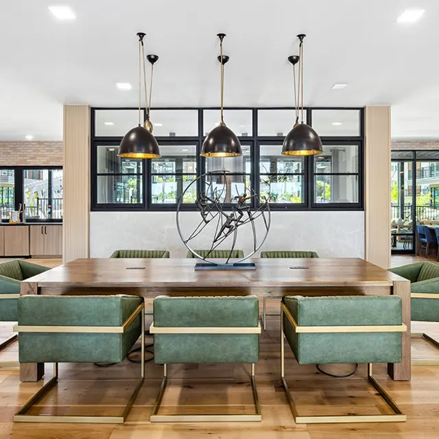 A modern dining area featuring a large wooden table surrounded by green upholstered chairs. Three hanging pendant lights illuminate the space, which has large windows allowing natural light in. A decorative sculpture is placed in the center of the table.