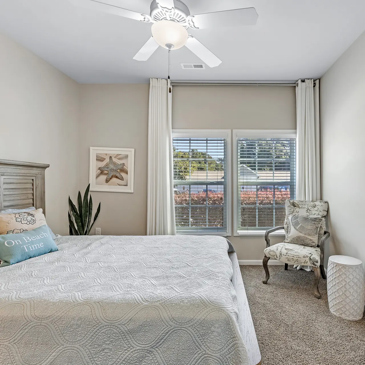 A well-decorated bedroom featuring a bed with decorative pillows, a bedside table, and large windows with white curtains letting in natural light.