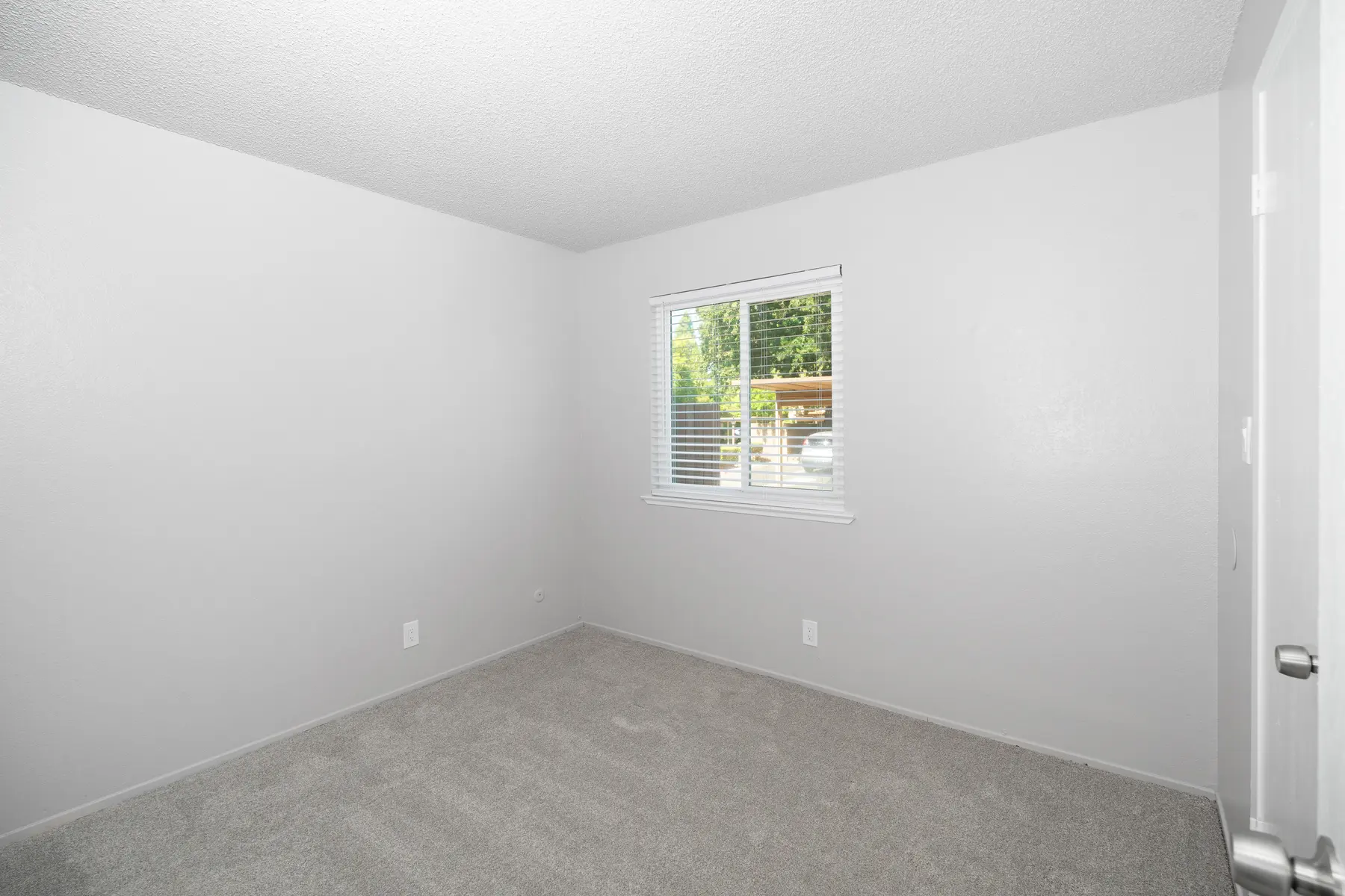 An empty room with gray walls and carpet, featuring a window with blinds that lets in natural light.