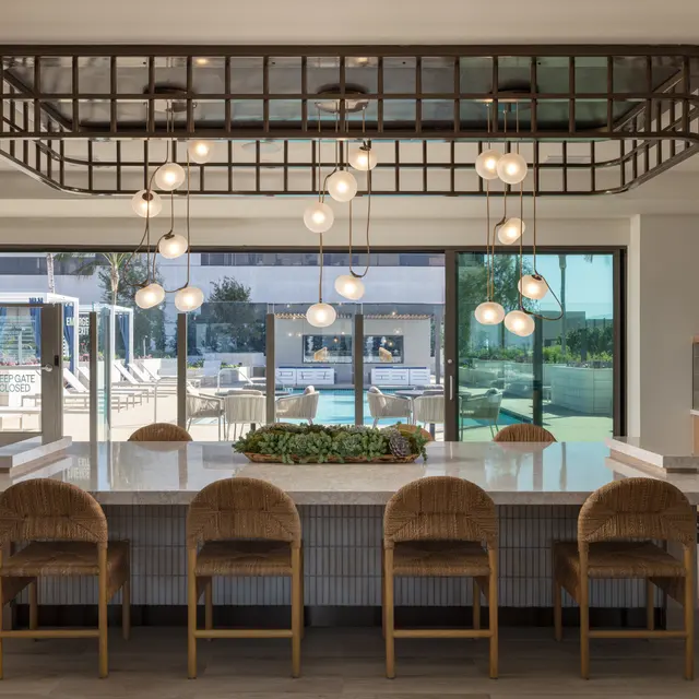 A modern kitchen featuring a large island with decorative plants, surrounded by stylish chairs. Unique light fixtures hang from a grid-like ceiling, creating an inviting dining space. Large sliding doors open to a view of an outdoor area with furniture.