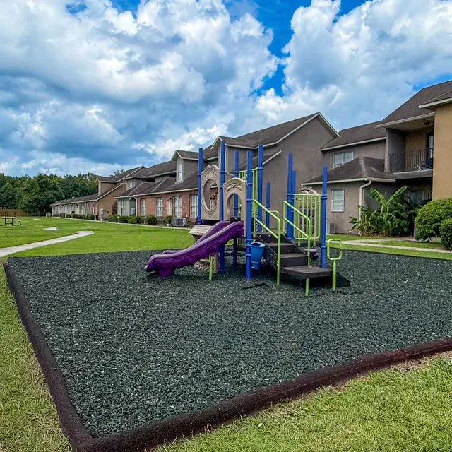 A colorful playground with climbing structures and a slide situated in a grassy area alongside residential buildings under a blue sky with clouds.