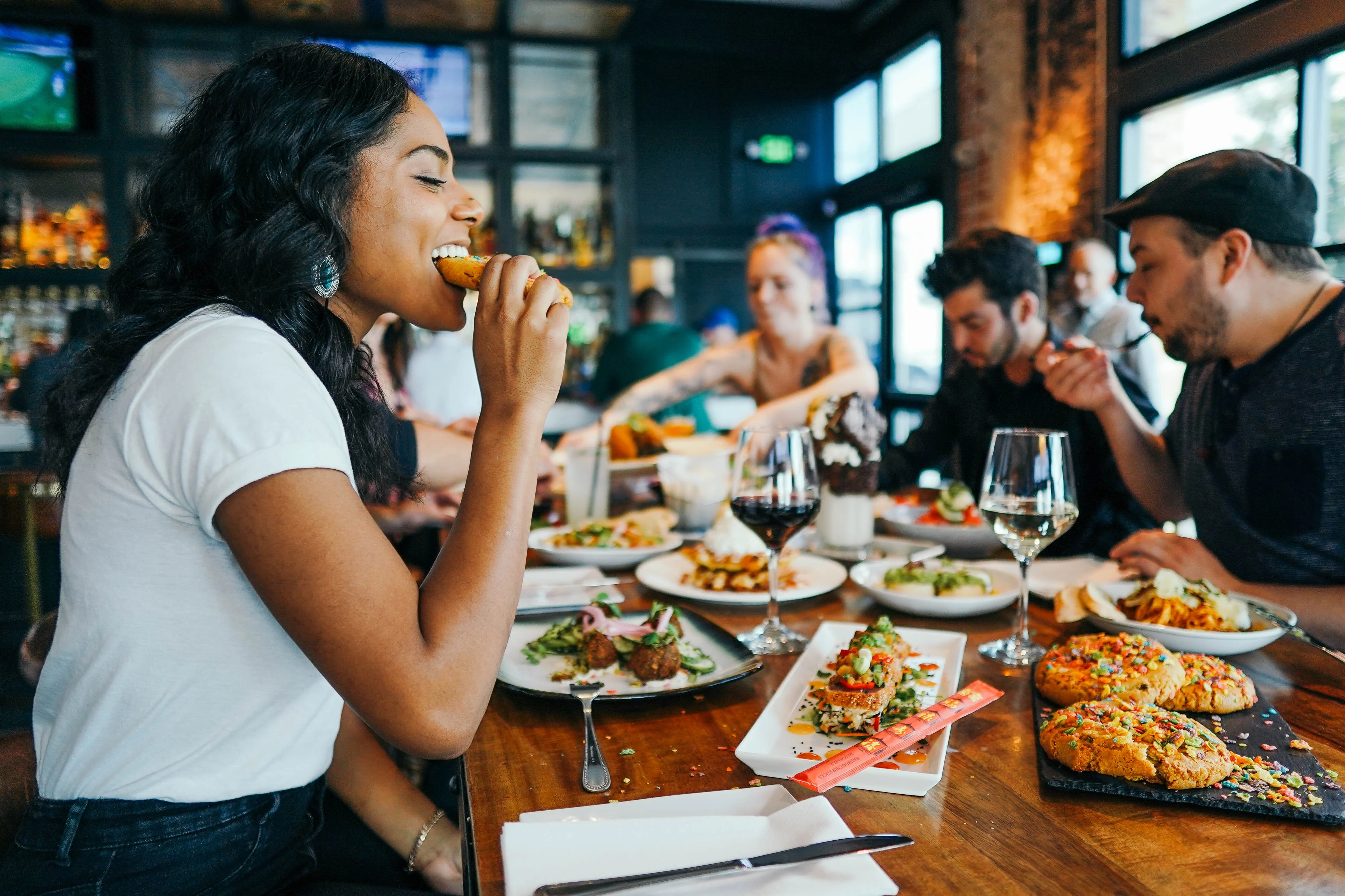 A group of friends enjoying a meal together at a restaurant with a variety of dishes on the table. One woman is taking a bite of her food, while others are engaged in conversation and eating.