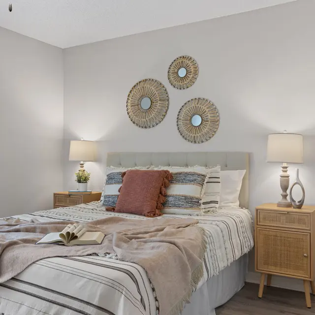 A cozy bedroom featuring a bed with layered bedding, decorative pillows, and a soft throw. The room includes bedside tables with lamps, decorative mirrors on the wall, and a warm color palette.