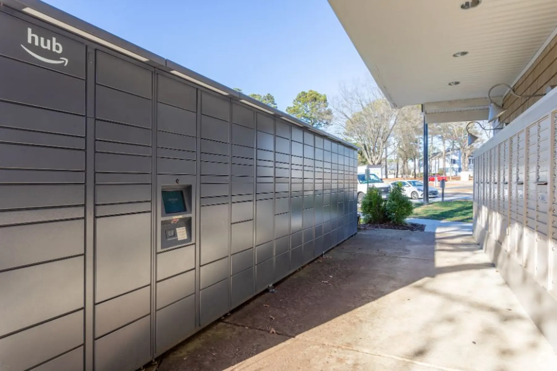 A modern package delivery locker system located outside a building, featuring a user interface for retrieving packages. The surrounding area includes green trees and a walkway.