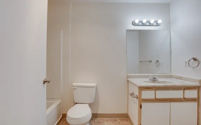 A clean and simple bathroom with a toilet, sink, and mirror. The walls are painted white, and there is a bathtub in the corner. Natural light floods the space.