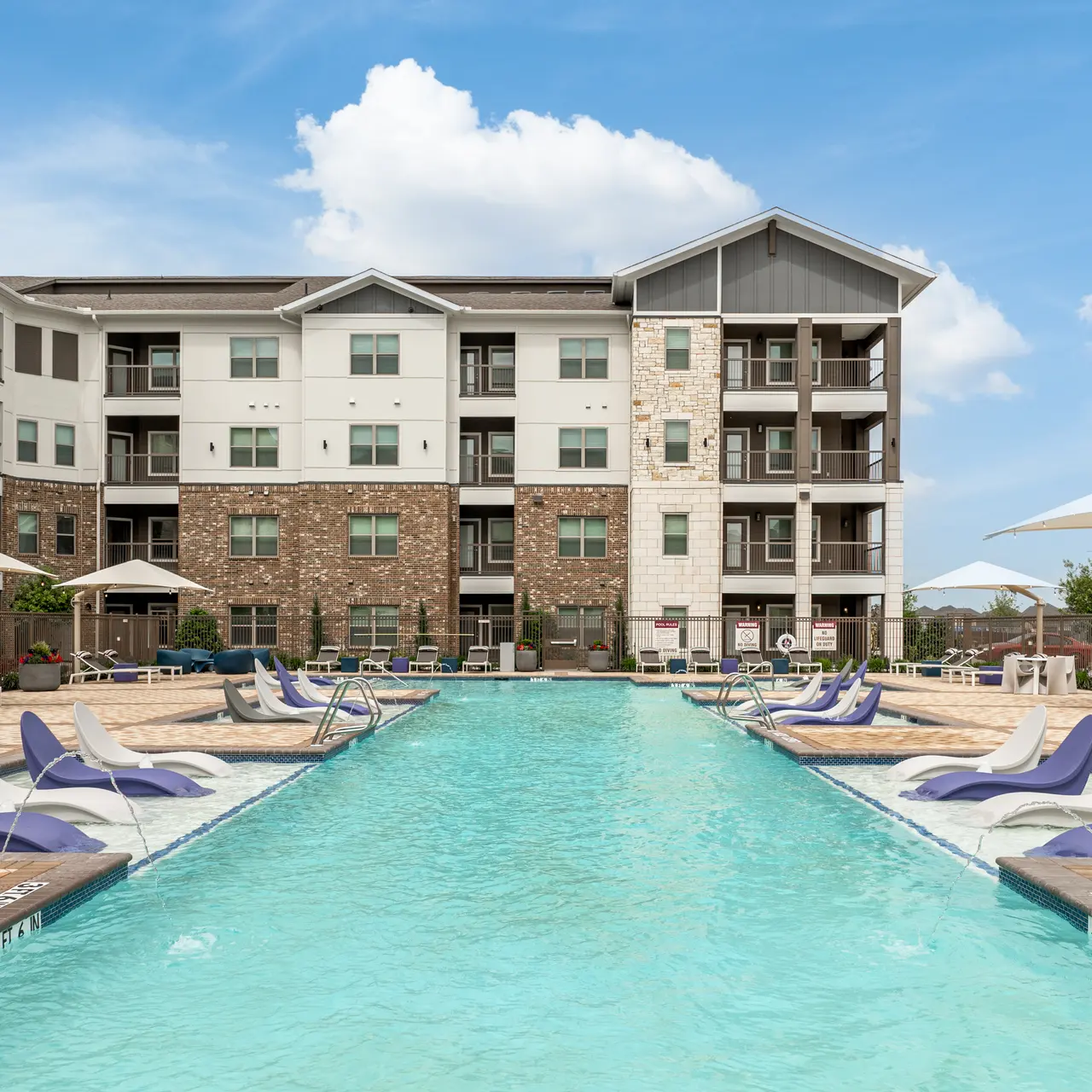 A modern swimming pool surrounded by lounge chairs and umbrellas, with an apartment building in the background under a clear blue sky.