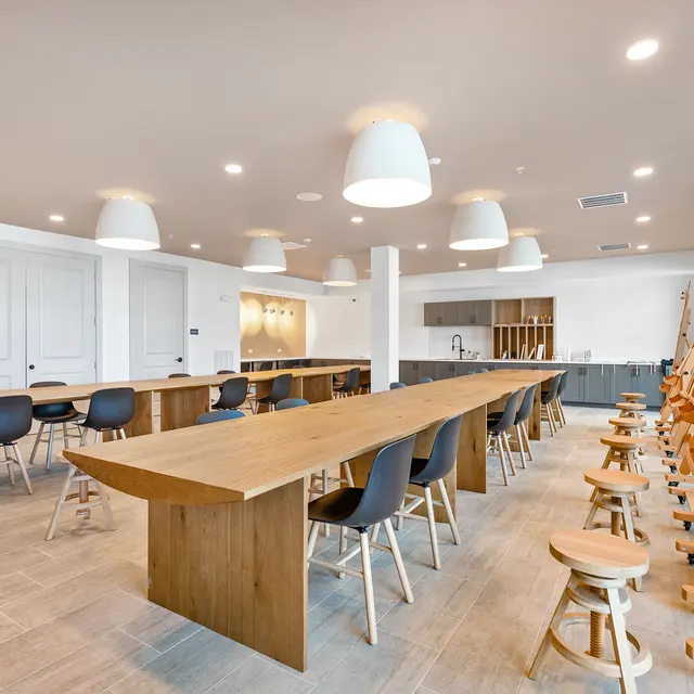A spacious art classroom featuring long wooden tables and stools. The room is well-lit with several pendant lights hanging from the ceiling. There are also easels positioned against the walls, indicating it is designed for artistic activities.