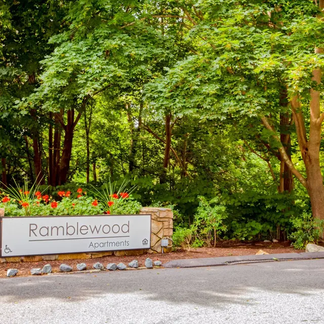 Sign for Ramblewood Apartments with a backdrop of lush green trees and flowers.