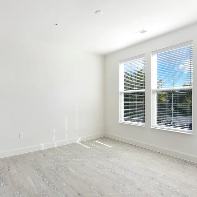 Spacious Empty Room An empty room with light-colored flooring and two large windows allowing natural light to enter. The walls are painted white, enhancing the bright and spacious feel of the room.