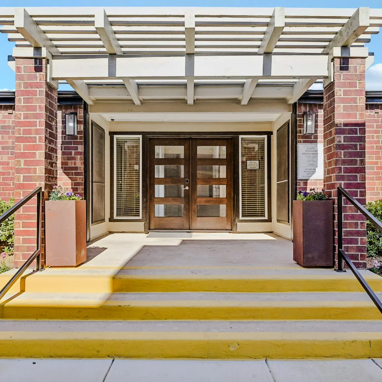 The entrance of a building featuring a covered porch, double glass doors, and yellow steps leading up to the door, flanked by potted plants.