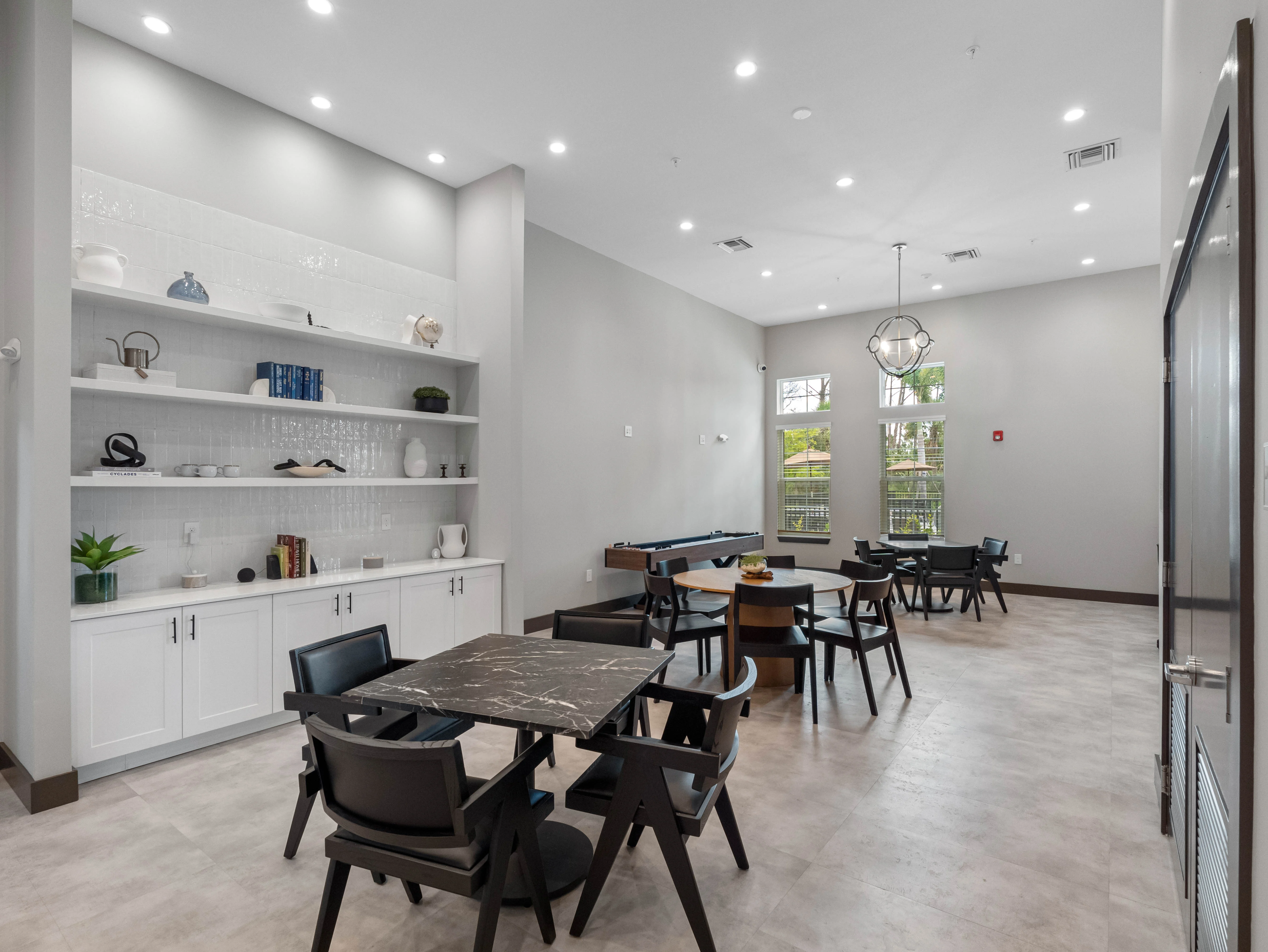 Contemporary Dining Area A stylish dining area featuring several tables and chairs arranged neatly. One wall has built-in shelves displaying decorative items. The room has bright overhead lighting and large windows allowing natural light.