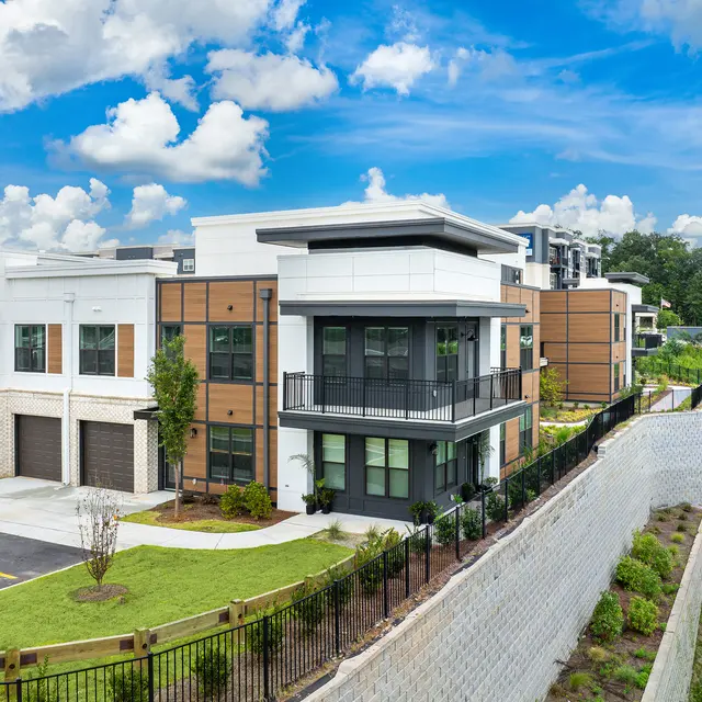 Modern Apartment Complex A modern apartment complex with a mix of white and brown wooden panels, featuring balconies and well-maintained landscaping. The sky is bright with fluffy clouds and the surrounding area has a green lawn and a stone wall.