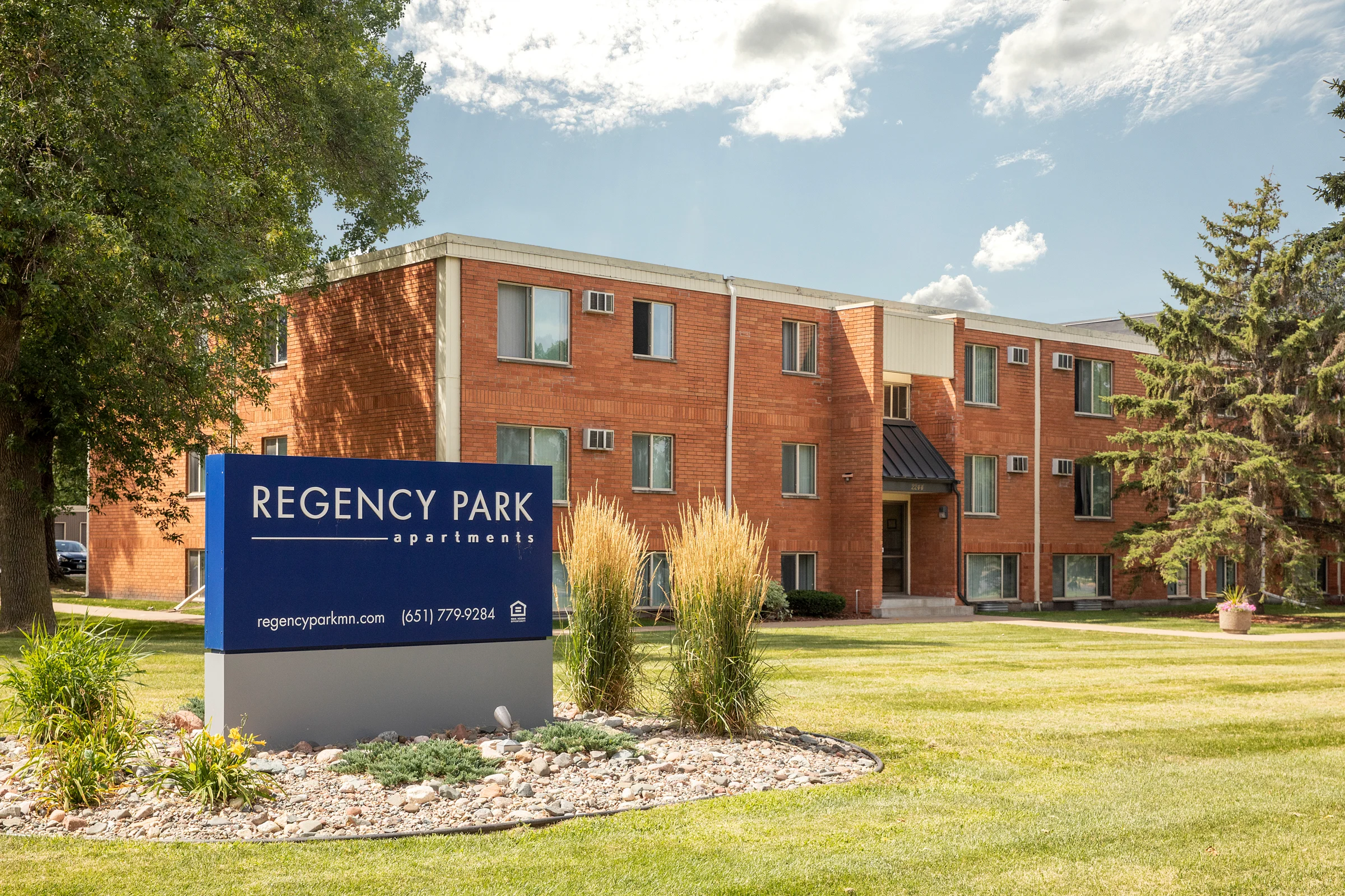 Regency Park Apartments Apartment complex with a sign that reads 'Regency Park Apartments'. The building is brick with windows and greenery around.