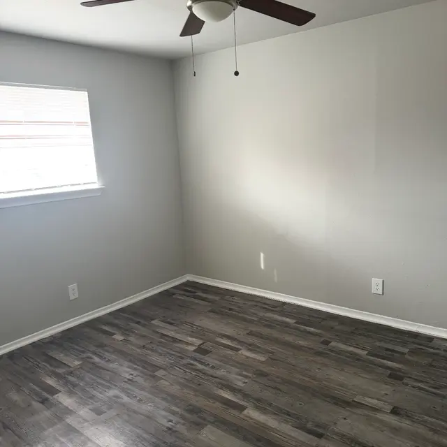 An empty room with a light gray wall, a window with blinds, and a ceiling fan. The floor is wooden with a dark finish.