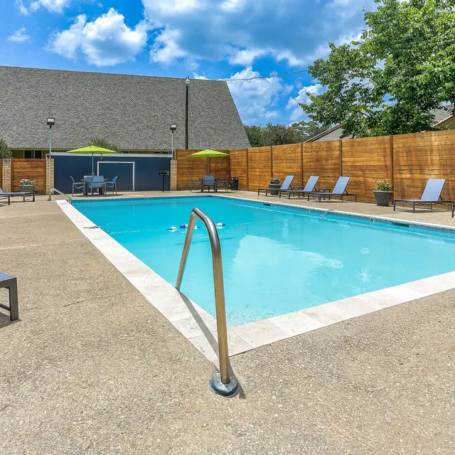 A bright, sunny swimming pool area surrounded by lounge chairs and umbrellas with a wooden fence and a building in the background.