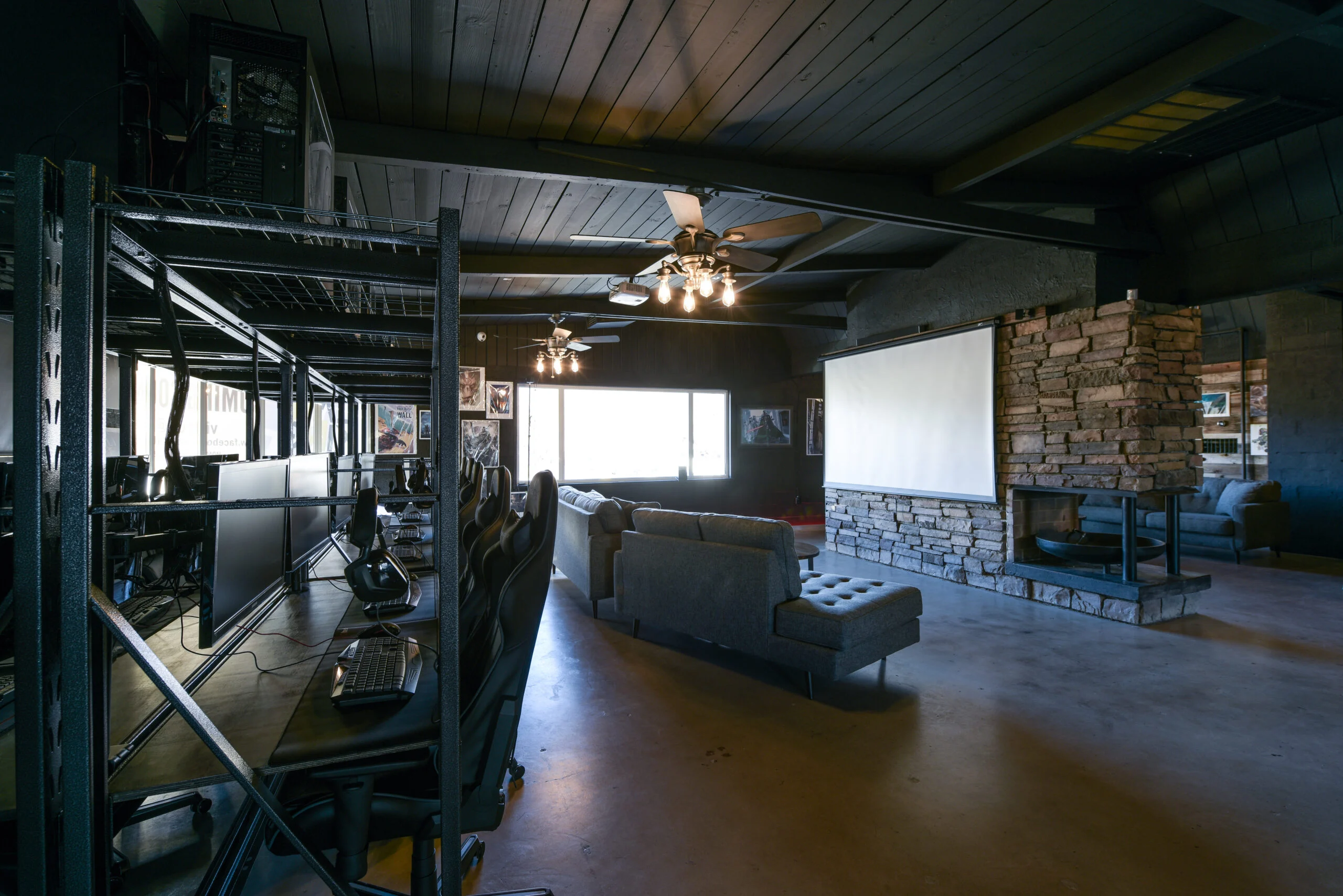Modern Living and Workspace A modern living space featuring a sectional couch, a stone fireplace, and several computer desks with monitors to the left. Large windows allow natural light to fill the room, and there is a projector screen on the wall opposite the couch.