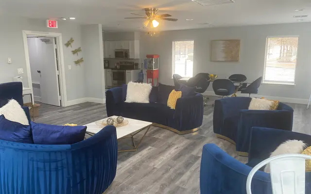 A modern living area featuring plush blue sofas, a coffee table, and a dining area with chairs in the background. The room is bright with natural light coming through the windows, and there are decorative elements like a painting on the wall and fluffy cushions on the sofas.