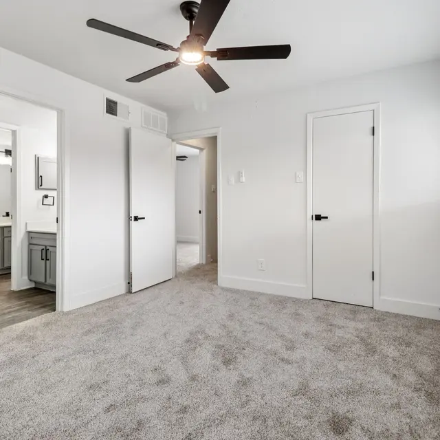 A spacious modern bedroom with light grey carpet, a ceiling fan, and white walls. Two open doors lead to adjacent rooms, and there's a window with blinds letting in natural light.