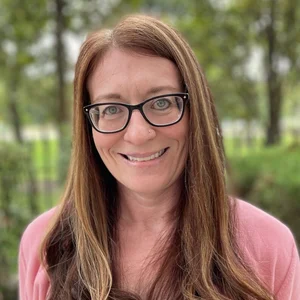 Outdoor Portrait of a Smiling Woman A woman with long brown hair and glasses smiles at the camera, standing outdoors with greenery in the background.