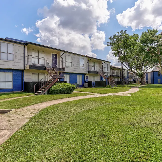 A view of an apartment complex with blue exteriors, featuring two-story buildings, a well-maintained lawn, and a pathway leading through the greenery.