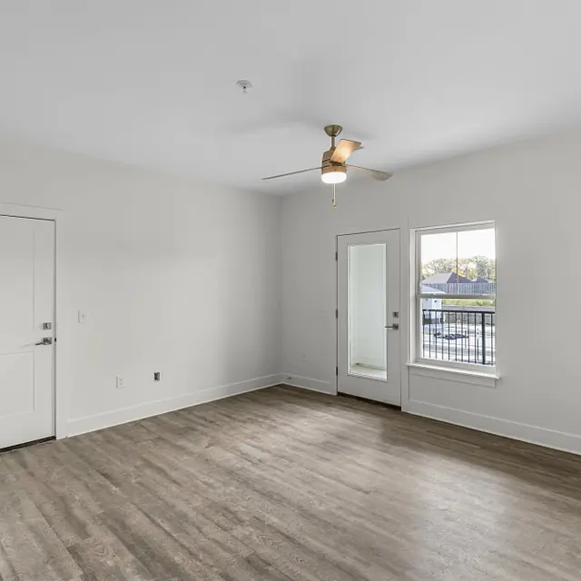 Spacious room in a modern apartment featuring a ceiling fan, light-colored walls, and large windows with a view of the outdoors.