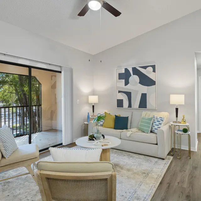 A stylish living room with a sofa adorned with colorful pillows, a coffee table, and a light rug. A sliding glass door leads to a balcony, with natural light flowing in from outside. Nearby, there's a hallway leading to other areas of the home.