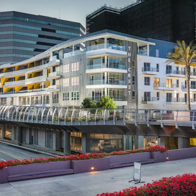 The Crest Residential Building A modern multi-story residential building with a white facade and green landscaping in front. The building features glass balconies and large windows, with palm trees visible nearby. In the background, there are taller skyscrapers and construction scaffolding.
